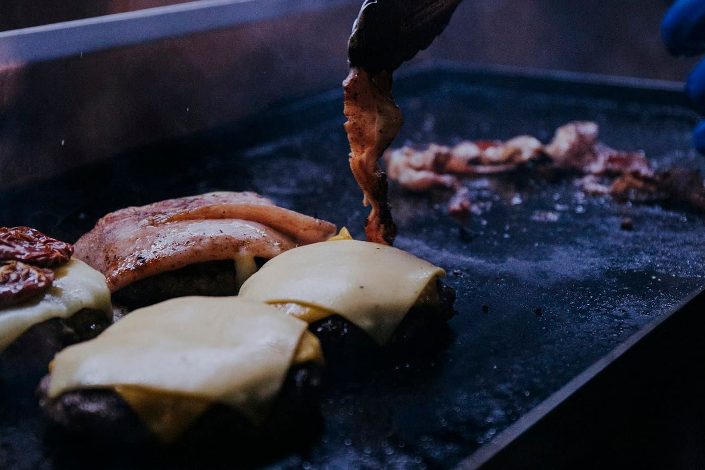 Chapa cheia de hambúrgueres com queijo no rush do evento, com a mão do operador virando o bacon.