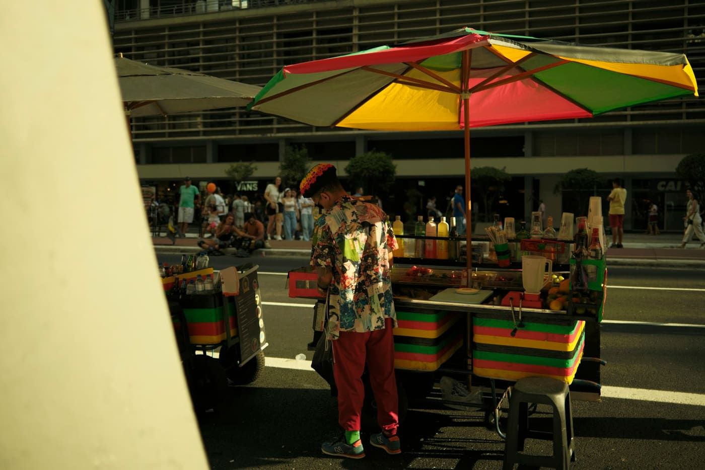 Expositor atendendo no carrinho sob guarda-sol colorido, na calçada de uma avenida de São Paulo.