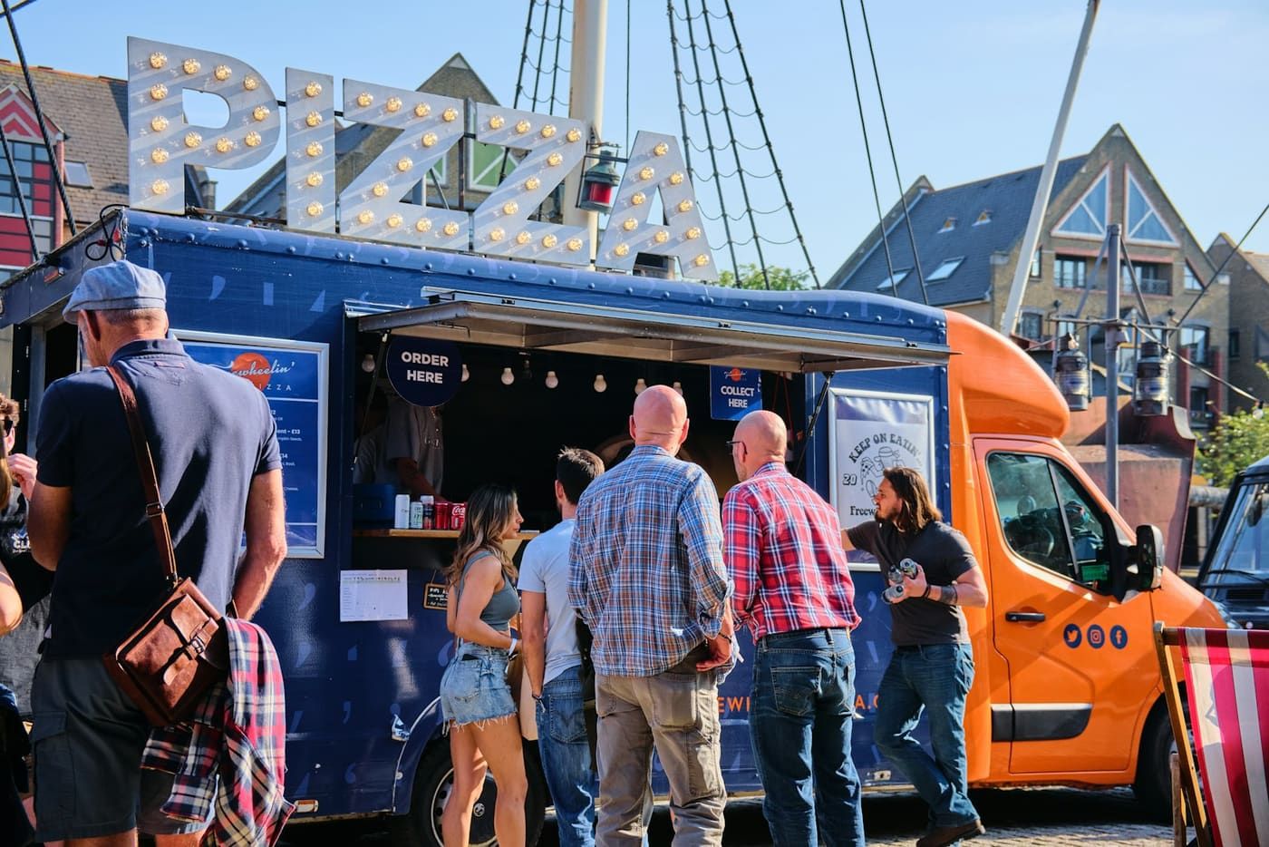 Fila de clientes no balcão de um food truck de pizza, num dia de sol.